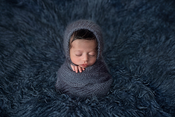 sweet image of a newborn baby boy wrapped in a blue knit wrap posed on a blue flokati rug at tanis saucier photography in her Montreal studio